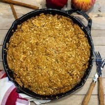 apple crisp in a cast iron skillet with cinnamon sticks and whole apples around the skillet